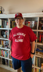 Brandon standing in front of a bookshelf wearing an "Alabama alumni" tshirt and an Alabama hat